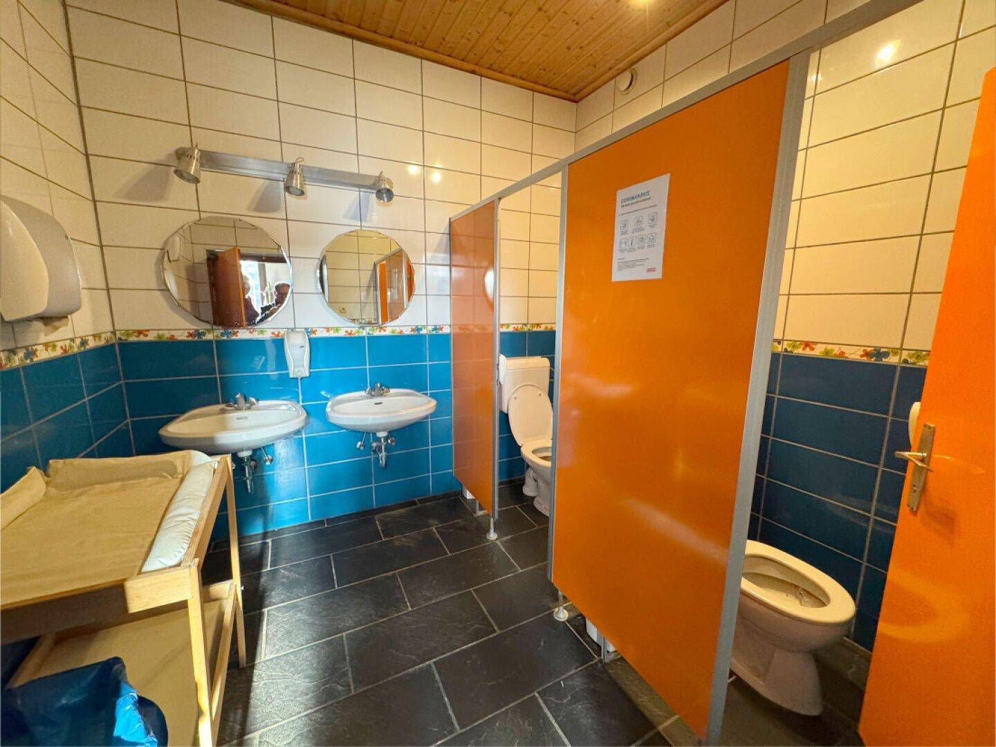 Öffentliche Toilette mit blauen und weißen Fliesen, Doppelwaschbecken und Wickeltisch.