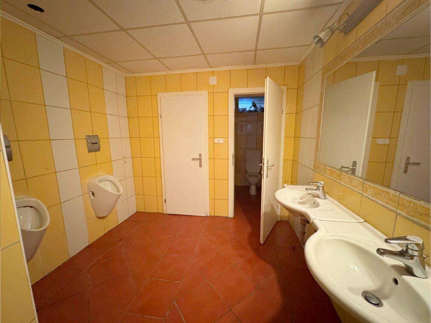 Helle öffentliche Toilette mit gelben Fliesen, Urinalen und zwei Waschbecken.