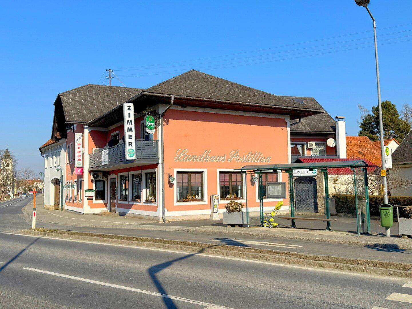 Seitenansicht des Landhauses mit Gastgarten und Bushaltestelle in ländlicher Umgebung.