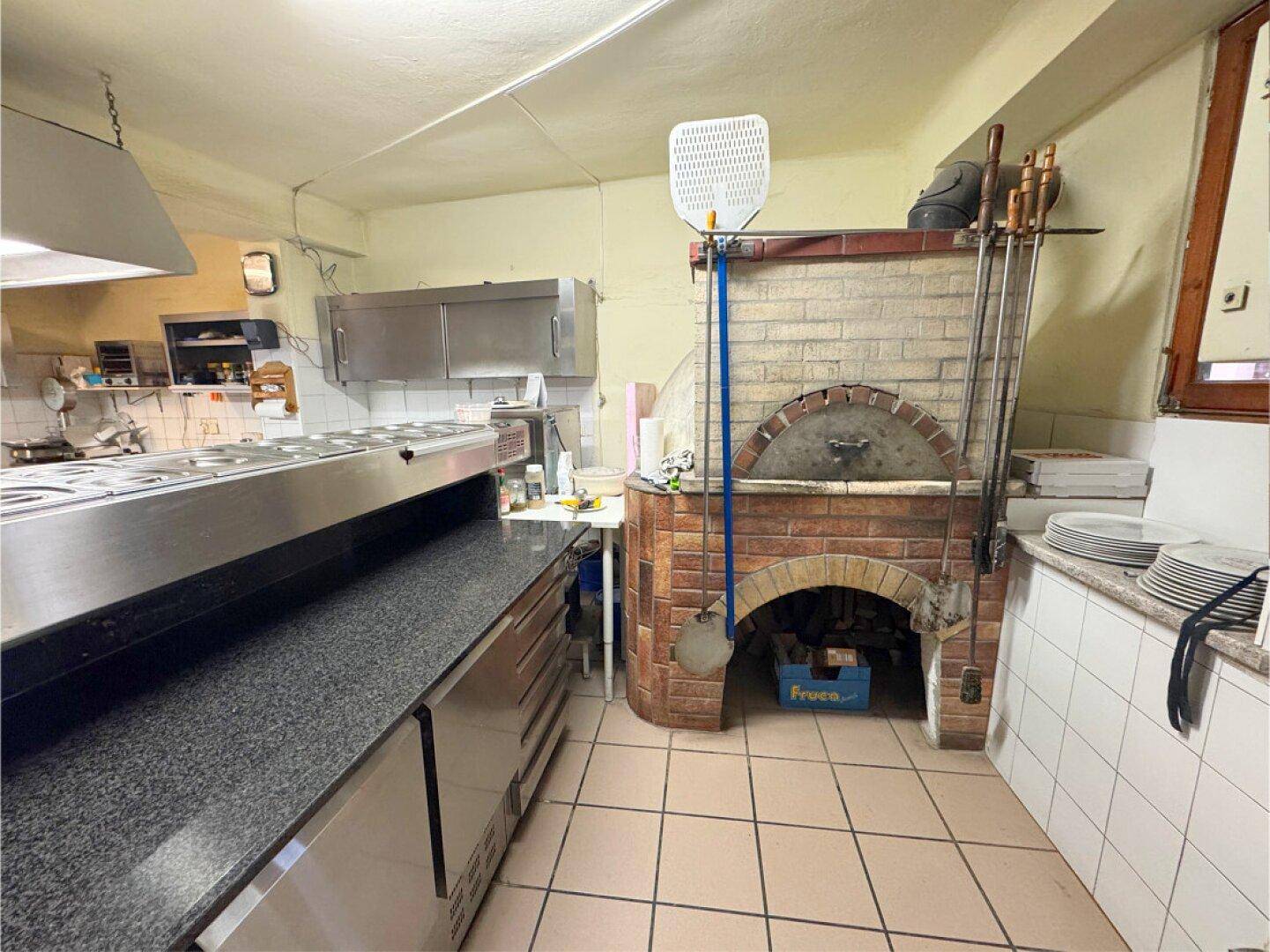 Professionelle Küche mit großem Pizzaofen, Edelstahlgeräten und Fliesenboden.