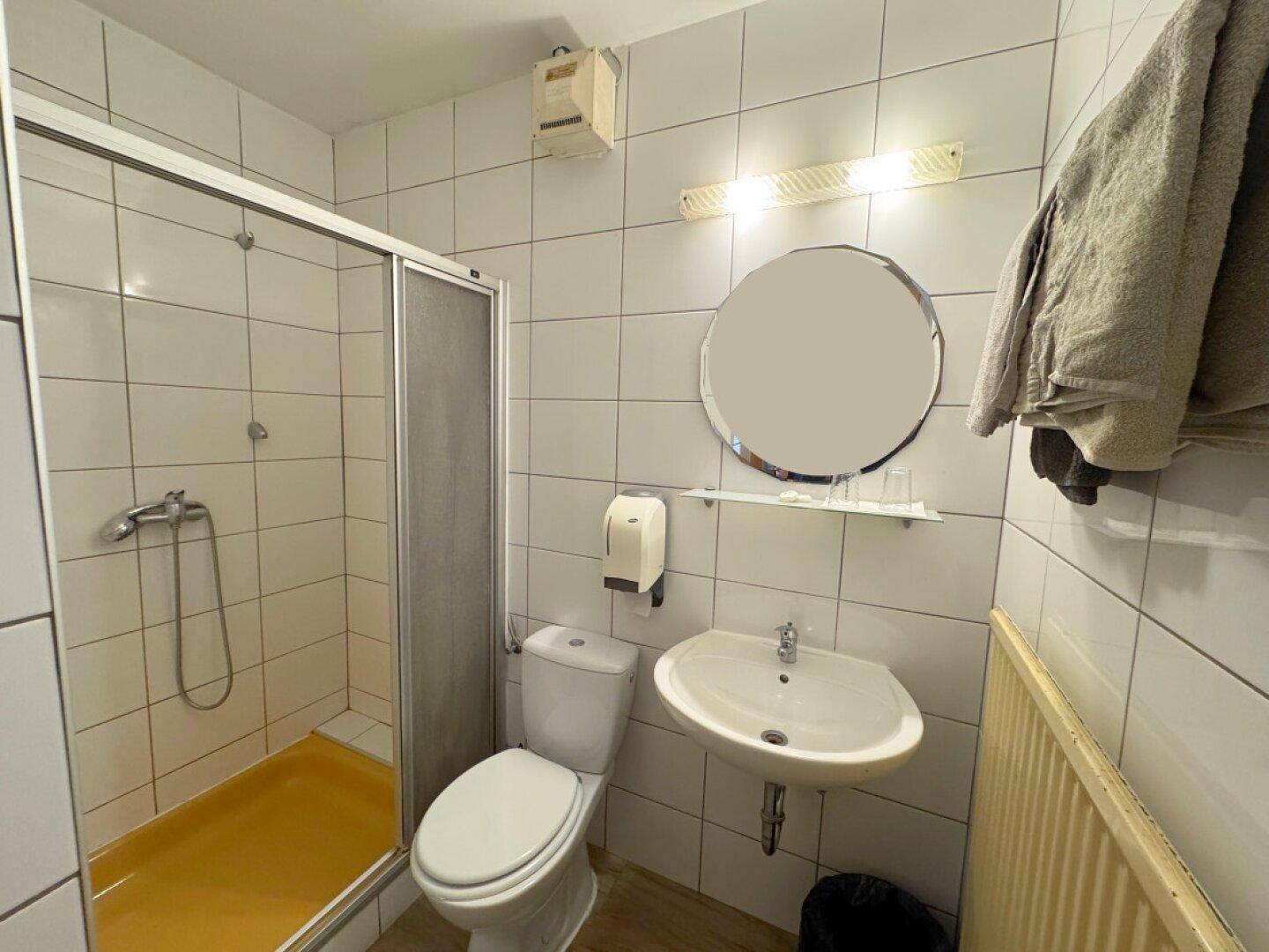 Einfaches Badezimmer mit Duschkabine, WC und Waschbecken in weiß gefliestem Raum.