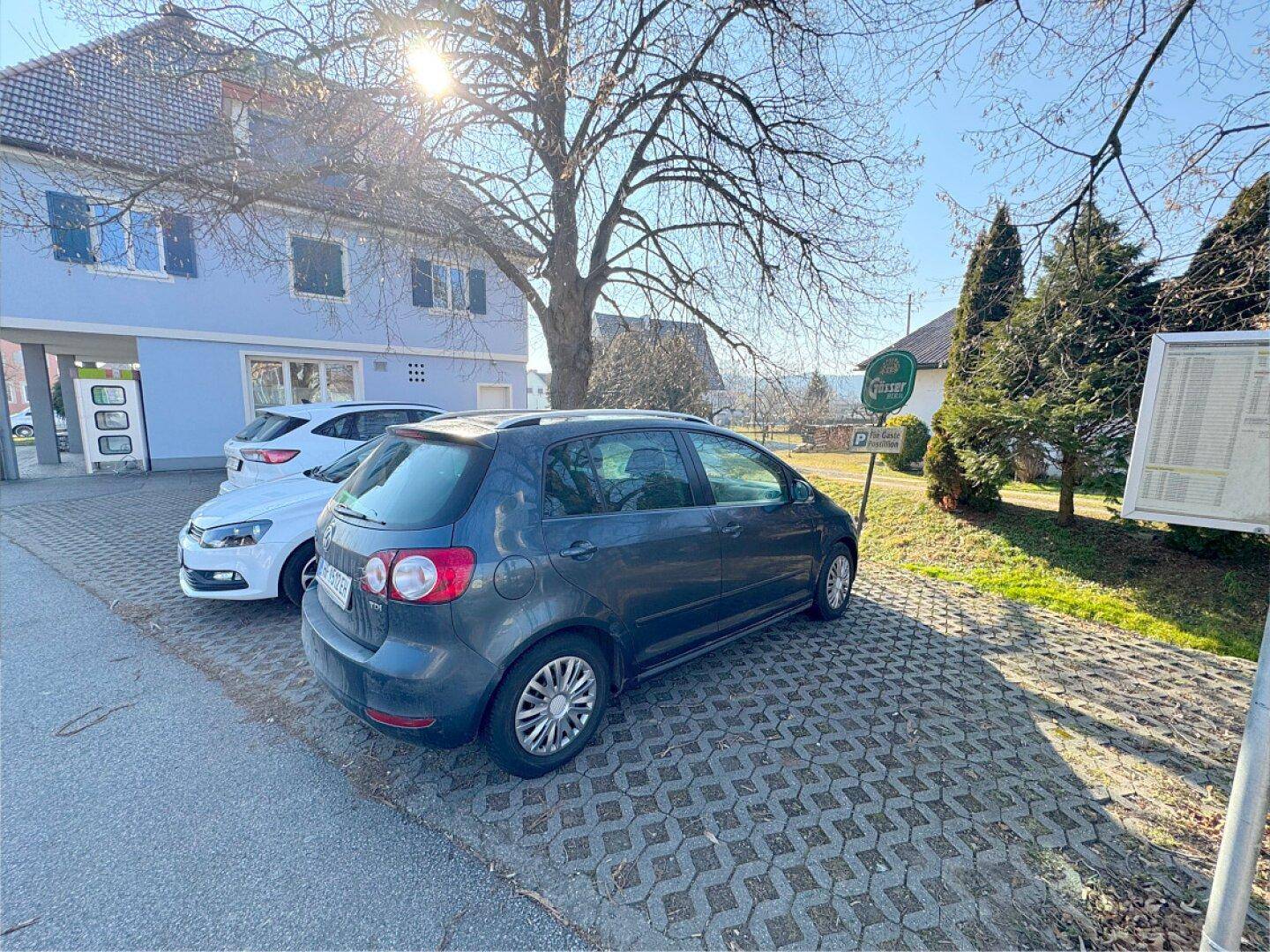 Parkplatz mit Pflastersteinen vor einem blauen Gebäude und Bäumen.