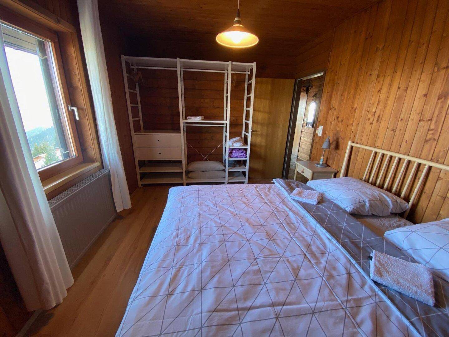 Schlafzimmer mit Holzboden, Holzvertäfelung, Doppelbett und offenem Kleiderschrank mit Blick ins Grüne.