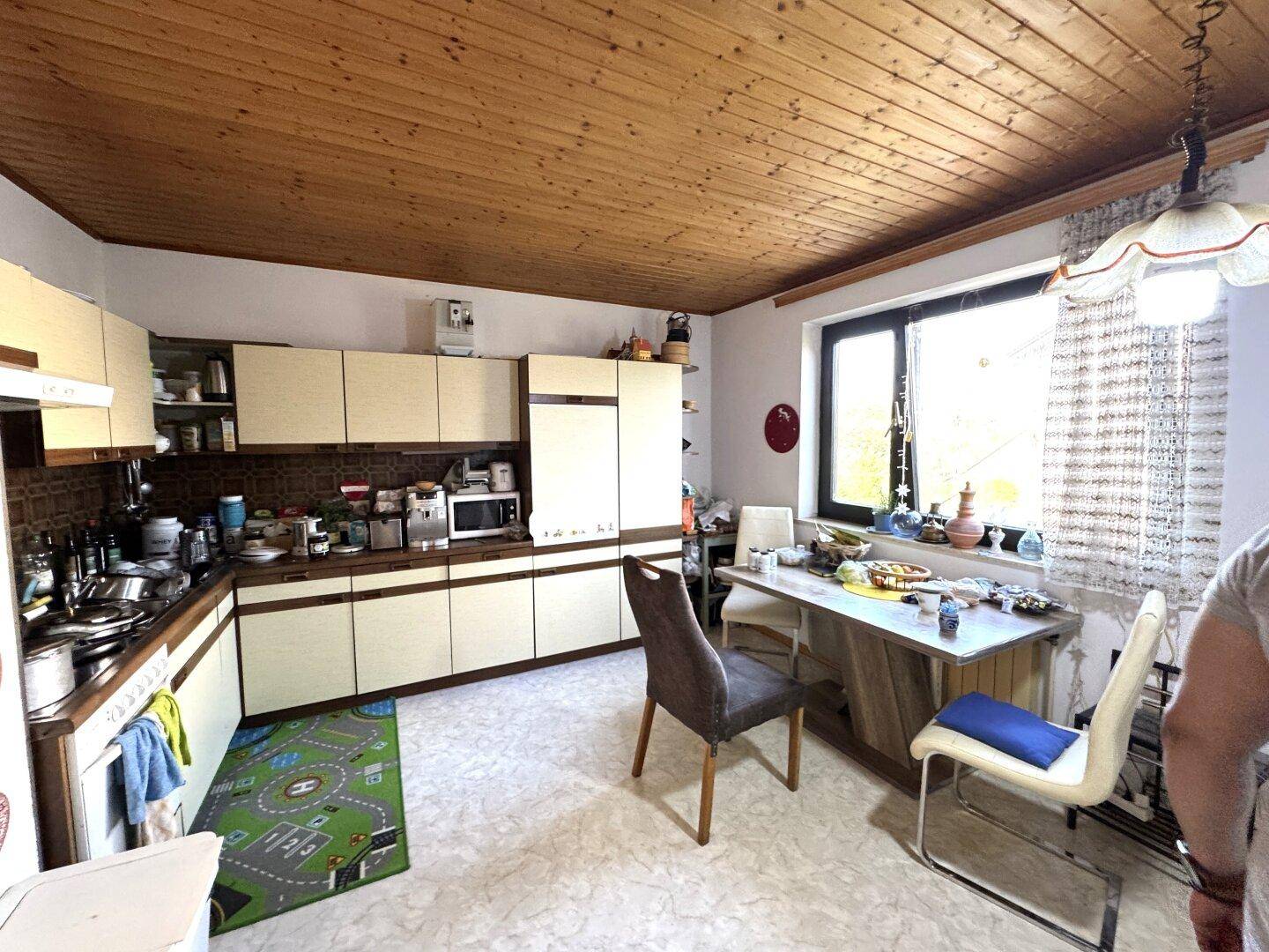 Küche mit Holzdecke, Einbauschränken und Essbereich am Fenster, ausgestattet mit älteren Geräten.