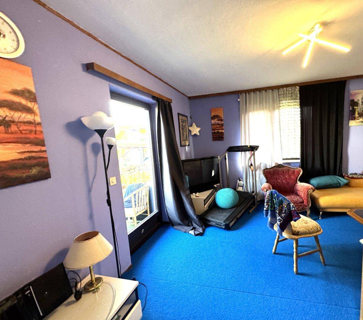 Buntes Wohnzimmer mit blauem Teppichboden, lila Wänden und Zugang zu einem Außenbereich.