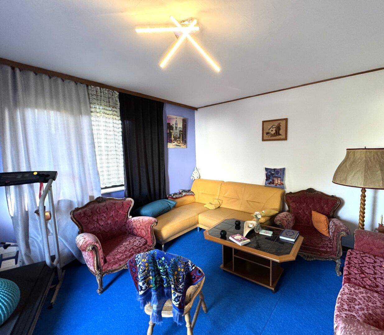 Wohnzimmer mit farbenfrohen Möbeln, blauem Teppichboden und einem Couchtisch in der Mitte.
