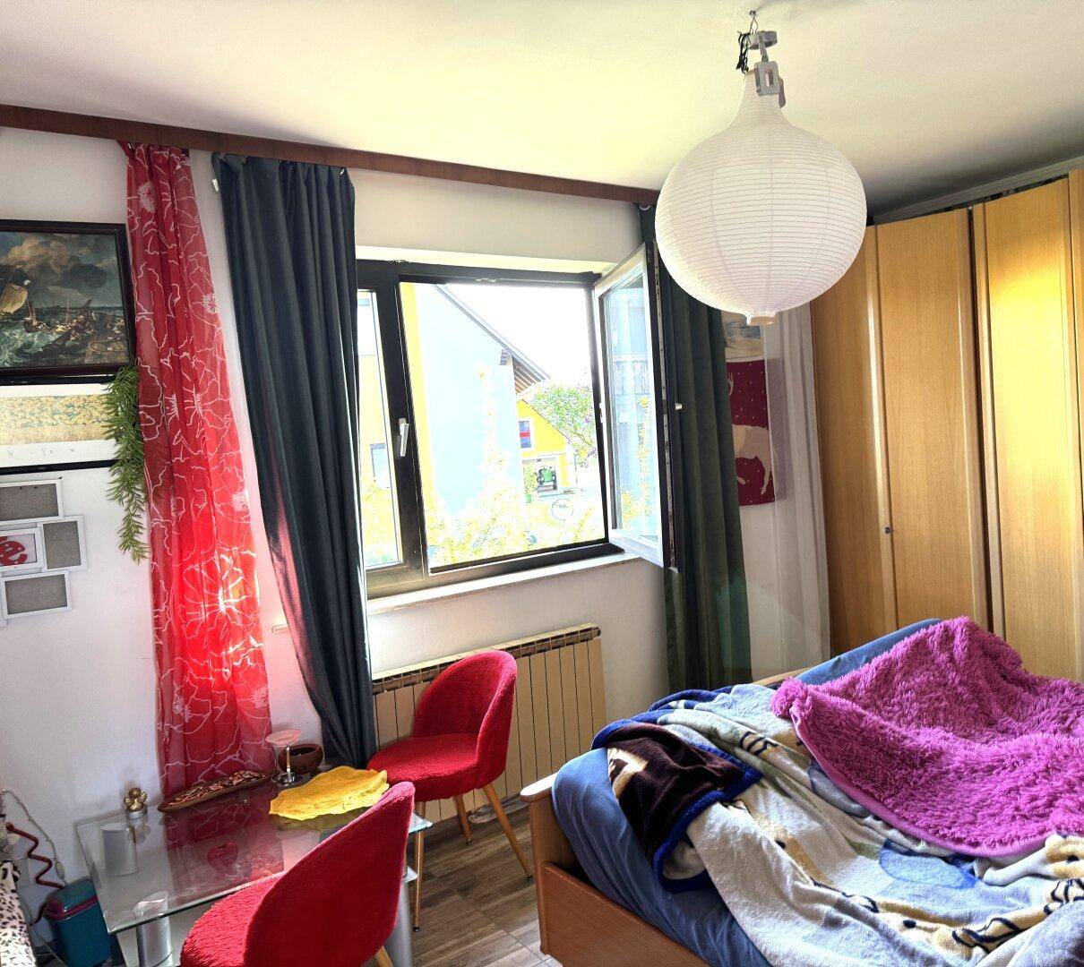 Schlafzimmer mit Bett, Kleiderschrank und Fenster, das viel Tageslicht hereinlässt.