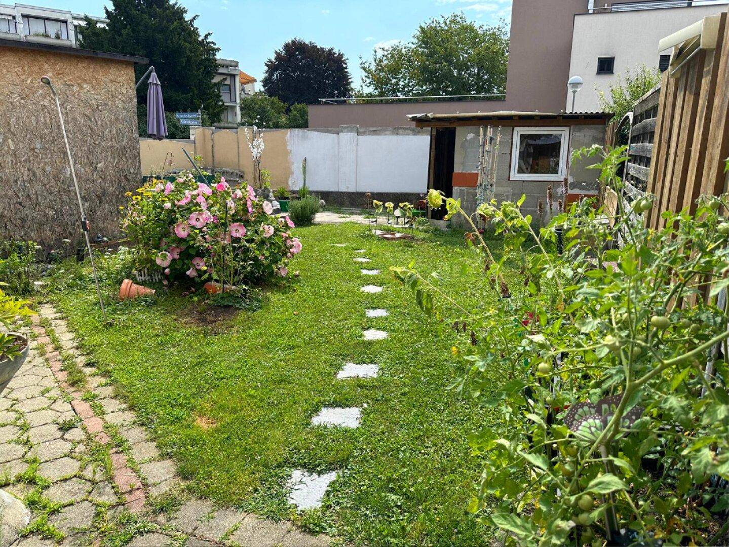 Grüner Gartenbereich mit Rasenfläche, Blumenbeeten und einem kleinen Gartenhaus im Hintergrund.
