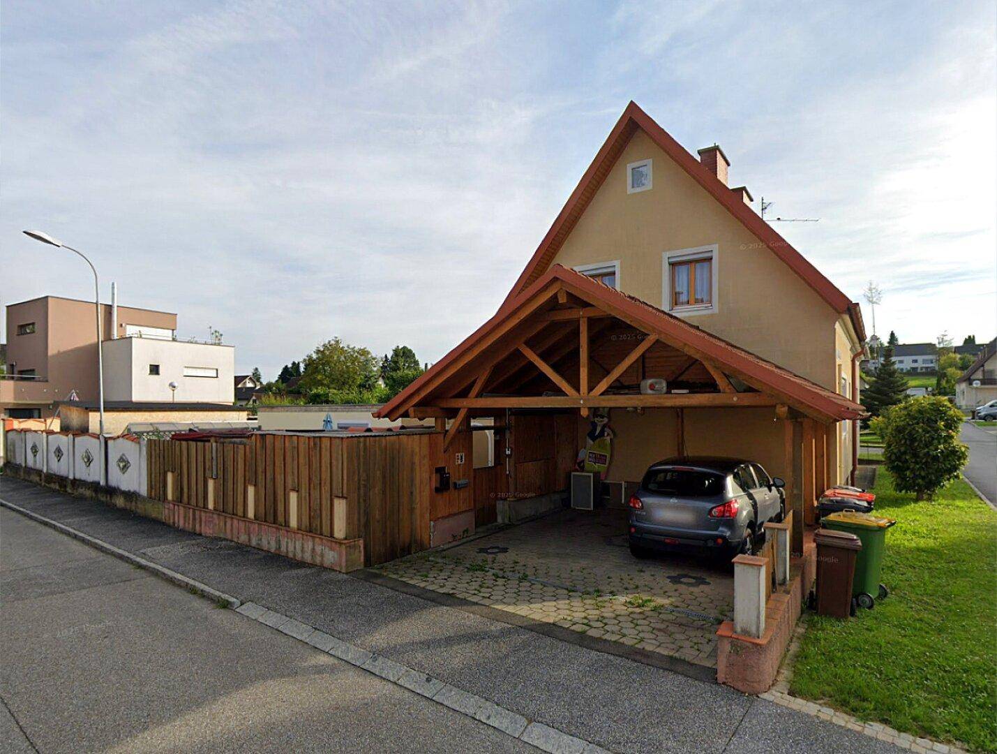 Wohnhaus mit Carport und kleiner Zufahrt, umgeben von einer Holzumzäunung in ländlicher Umgebung.