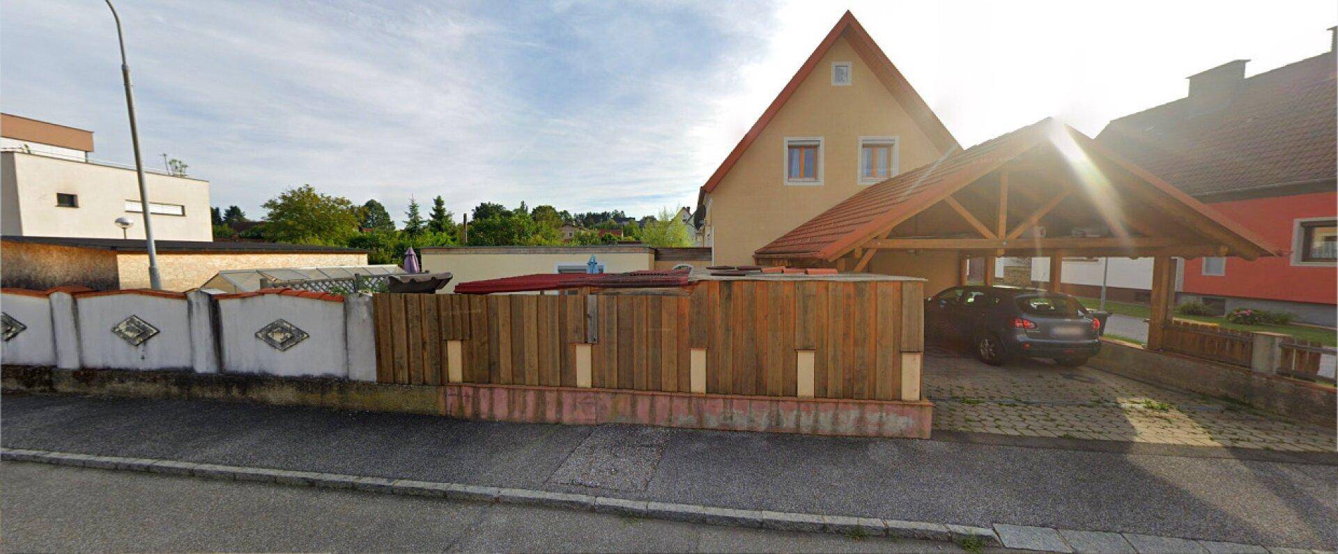 Einfamilienhaus mit Carport und sonniger Ausrichtung, umgeben von einer Holzumzäunung am Straßenrand.