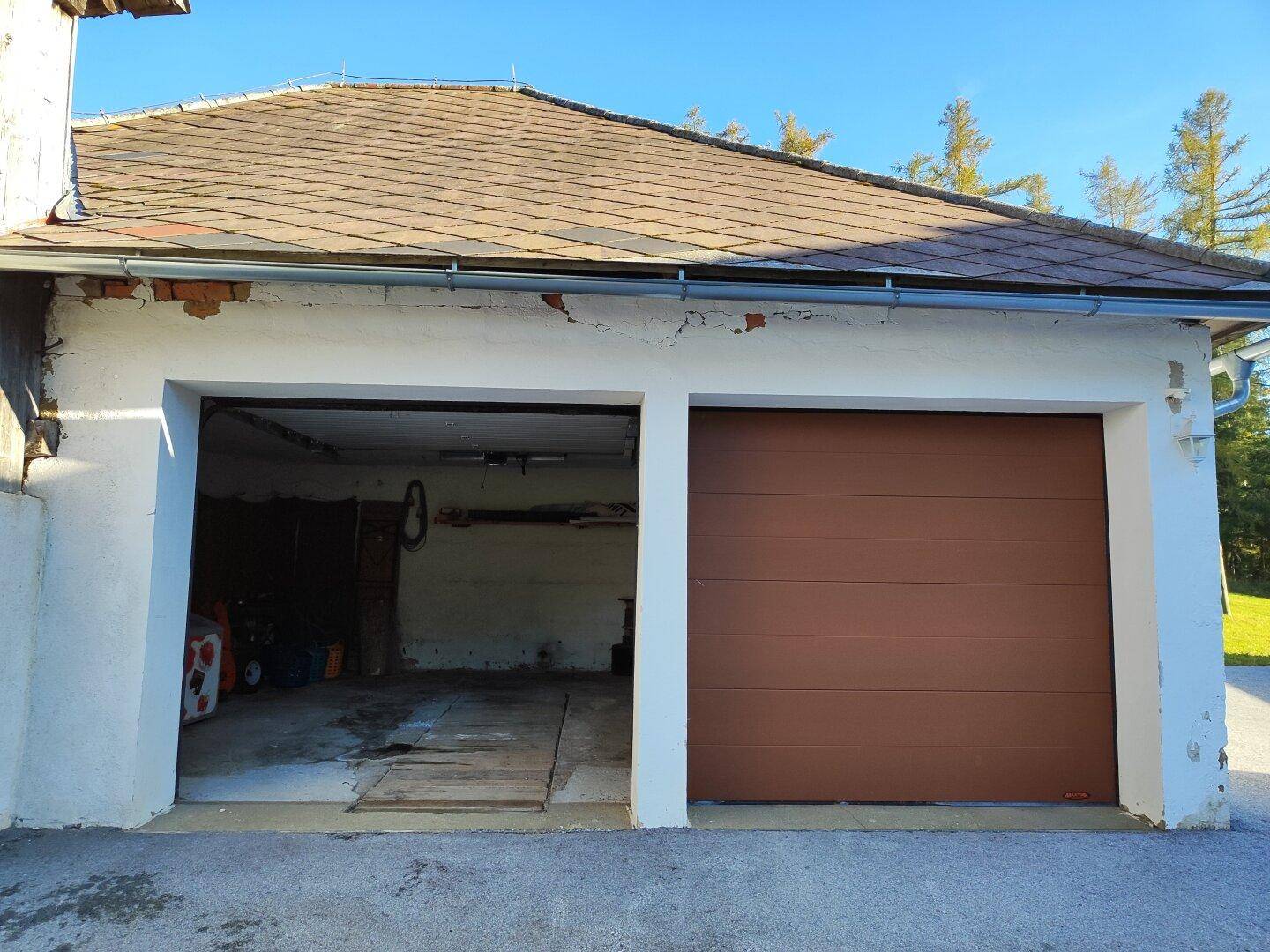 Außenansicht einer Garage mit zwei Toren, eines davon geöffnet, unter einem traditionellen Ziegeldach.