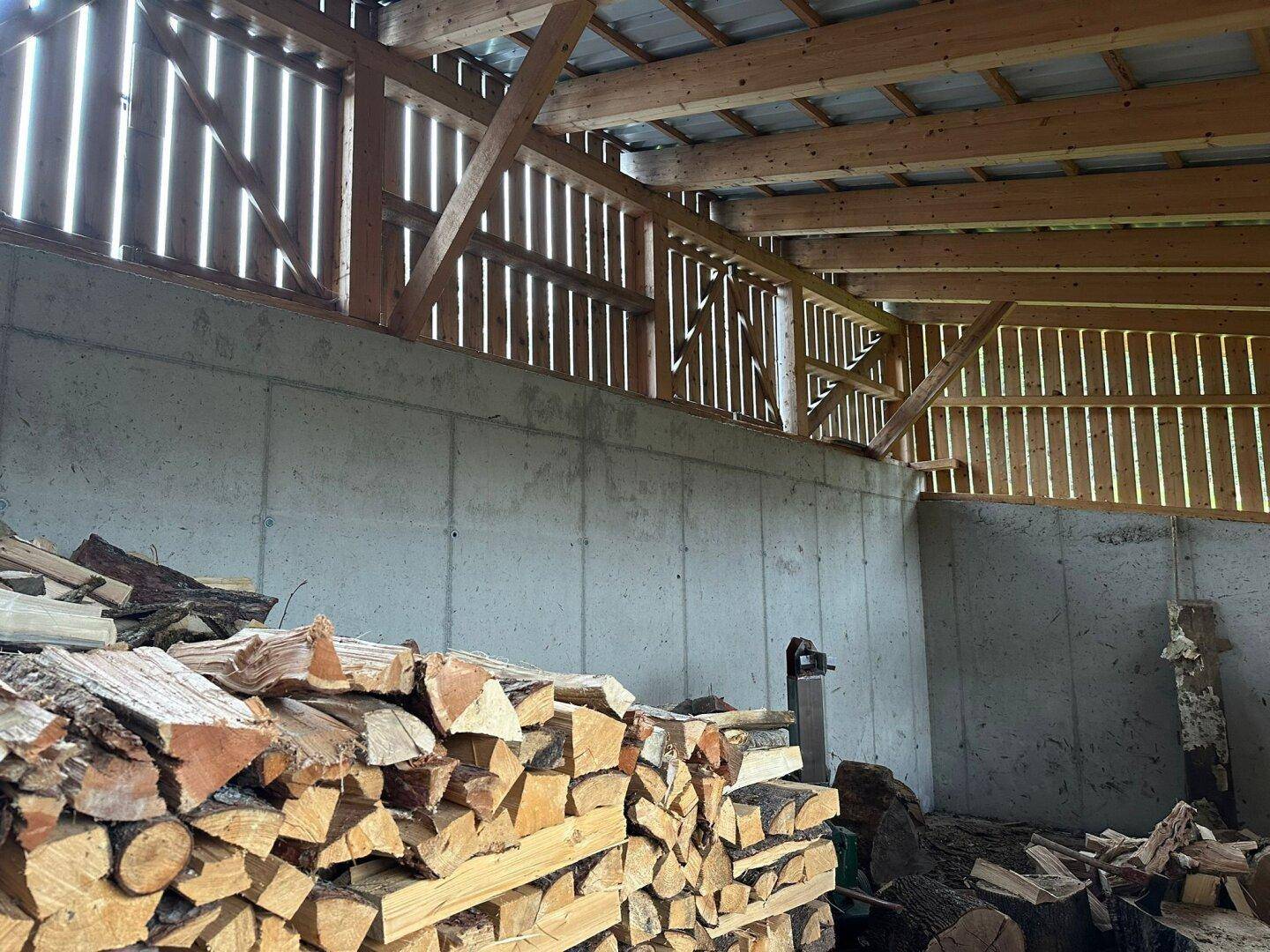 Innenraum einer Scheune oder eines Lagerraums mit gestapeltem Brennholz und sichtbaren Holzbalken.