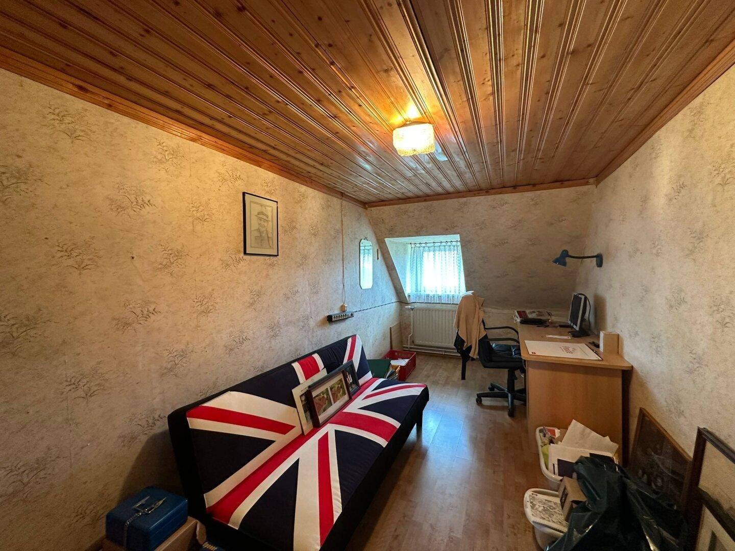 Zimmer mit Sofa im Union-Jack-Design, Schreibtisch und Holzdecke, hell durch ein Fenster.