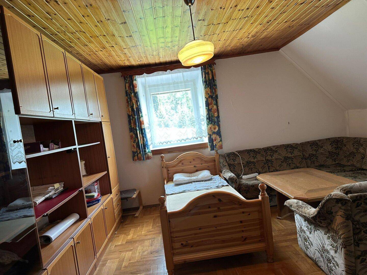 Gemütliches Schlafzimmer mit Einzelbett, Sofa und Holzdecke, hell durch ein Fenster.
