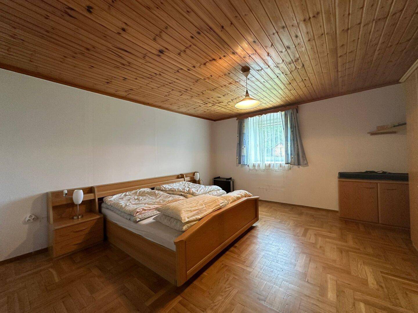 Schlafzimmer mit Doppelbett, Holzdecke und Fischgrätparkett, hell durch ein Fenster.