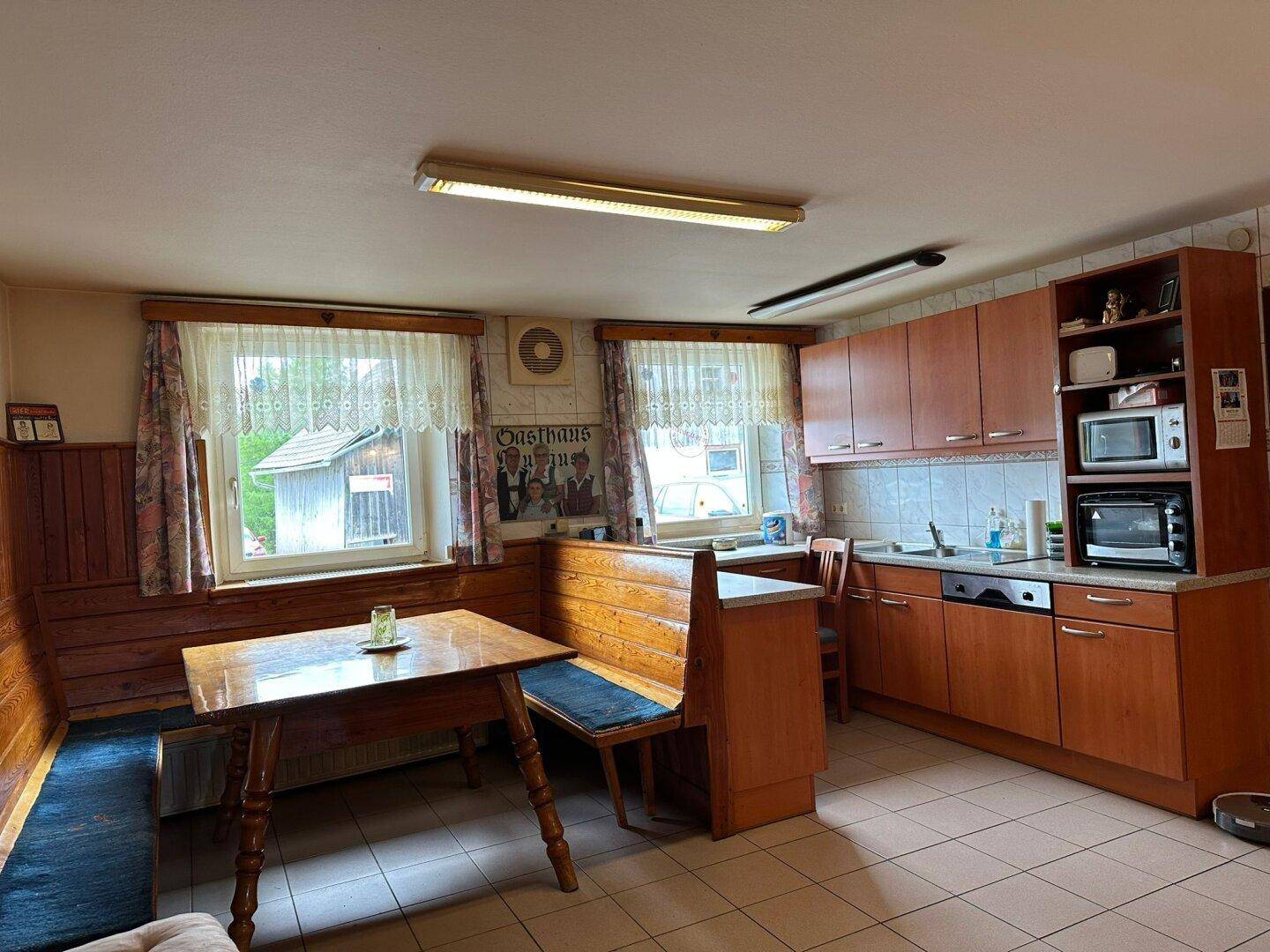 Funktionale Küche mit Holzschränken, Essbereich und Fliesenboden, hell durch Fenster.