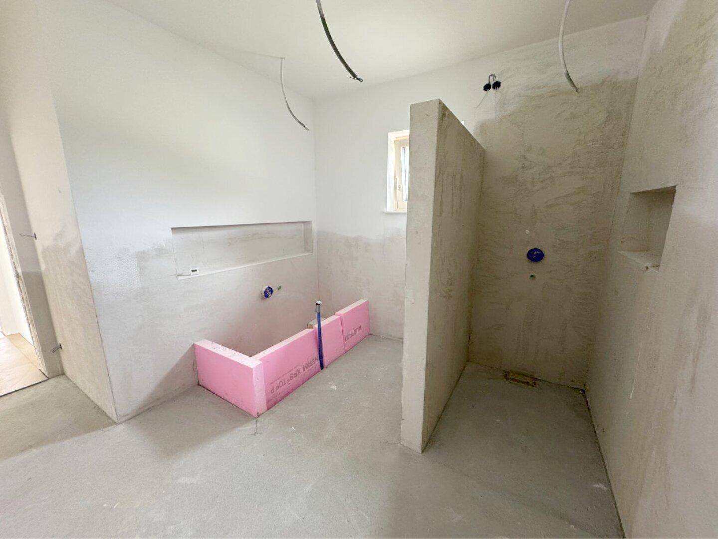 Geräumiges Badezimmer im Rohbau mit Fenster und vorbereitetem Bereich für eine Dusche.