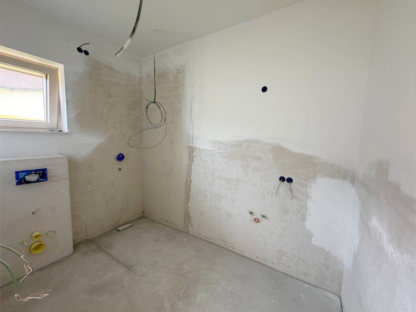 Badezimmer im Rohbau mit Fenster und vorbereiteten Anschlüssen für Sanitärinstallationen.