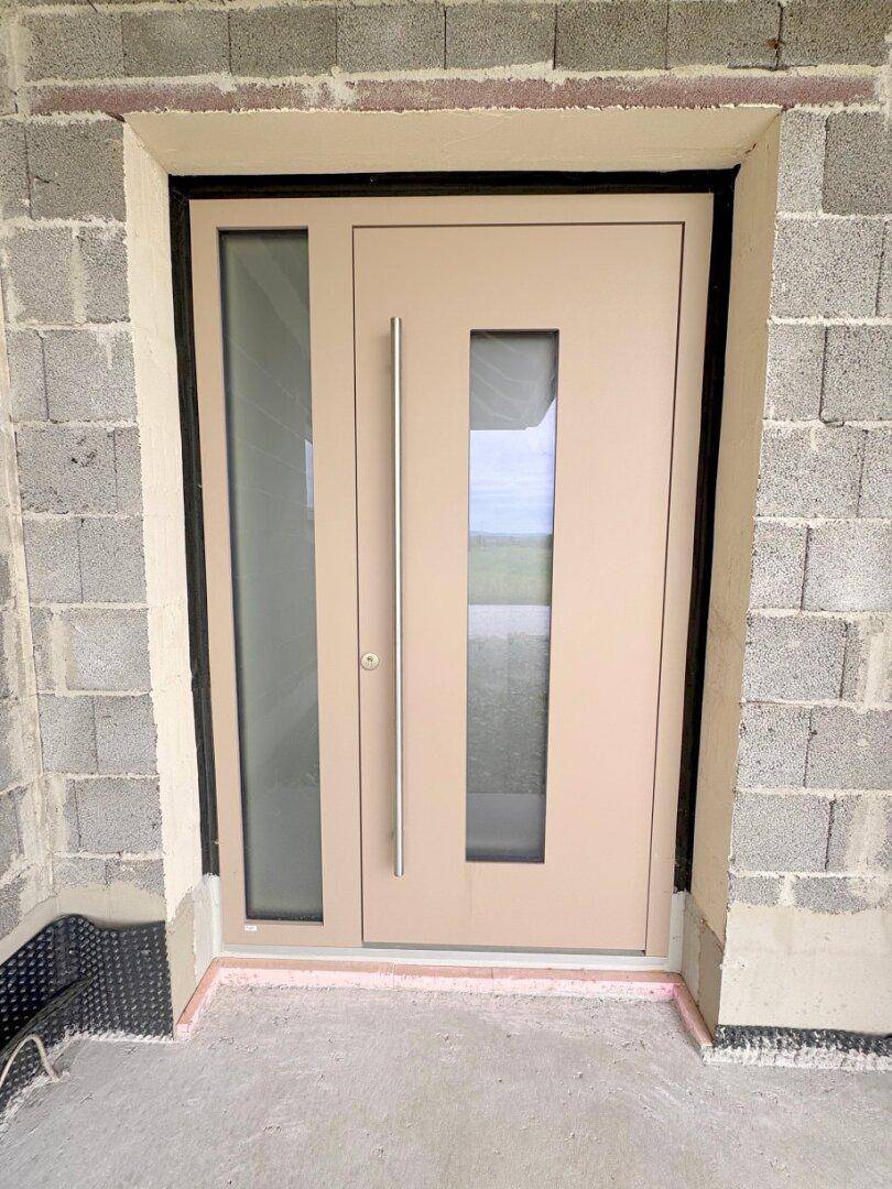 Moderne Eingangstür in Beige mit Glaseinsätzen und langem Griff, noch im Rohbau.
