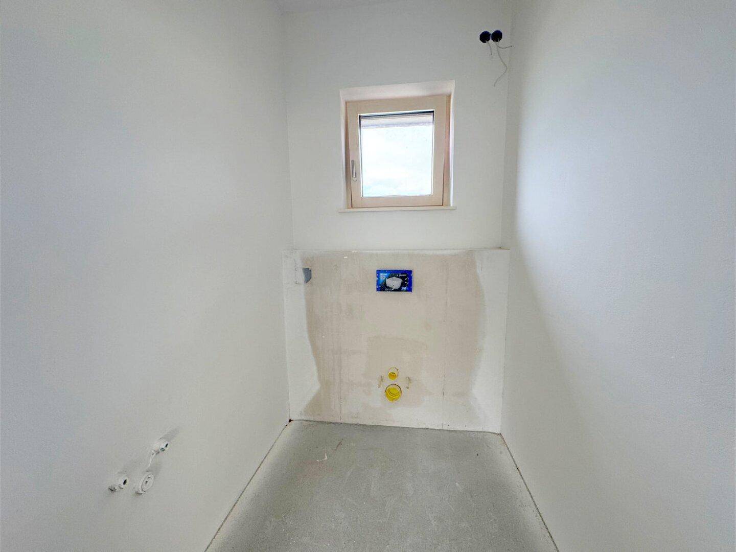 Kleines WC im Rohbau mit Fenster und vorbereiteten Anschlüssen für die Toilette.