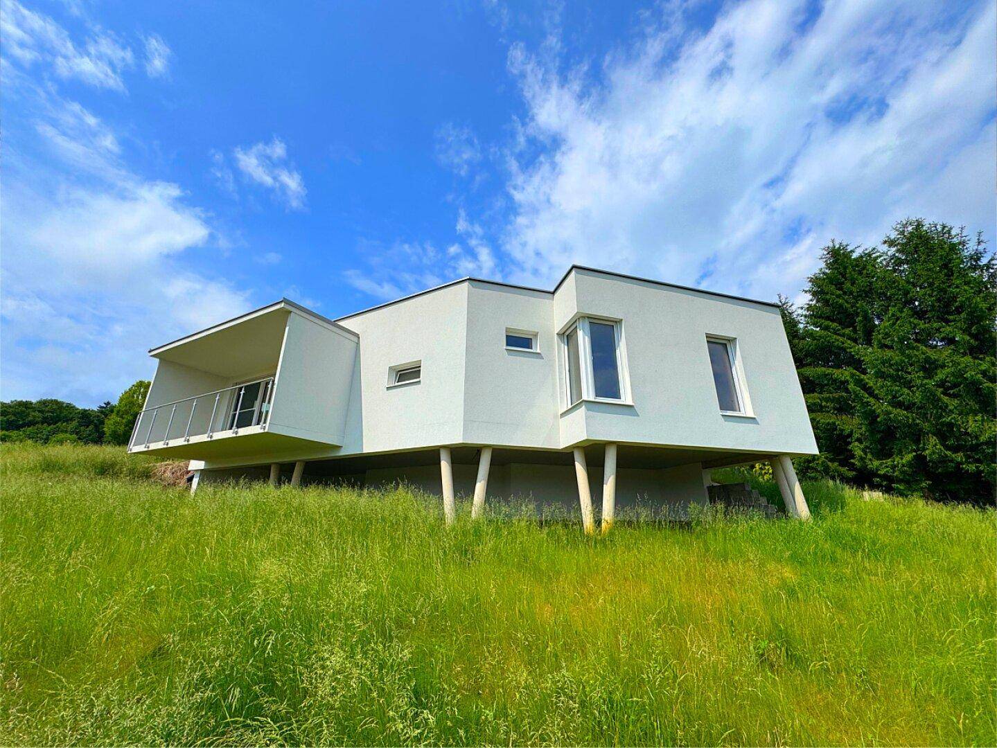 Zeitgenössisches weißes Haus mit Flachdach und Balkon, eingebettet in eine grüne Landschaft.