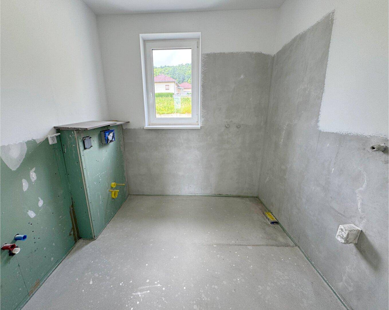 Unfertiges Badezimmer mit Fenster und vorbereiteten Anschlüssen für Sanitärinstallationen.