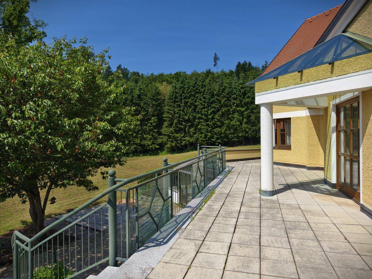 Geräumige geflieste Terrasse am Haus mit Blick auf die umliegende grüne Landschaft und Bäume.