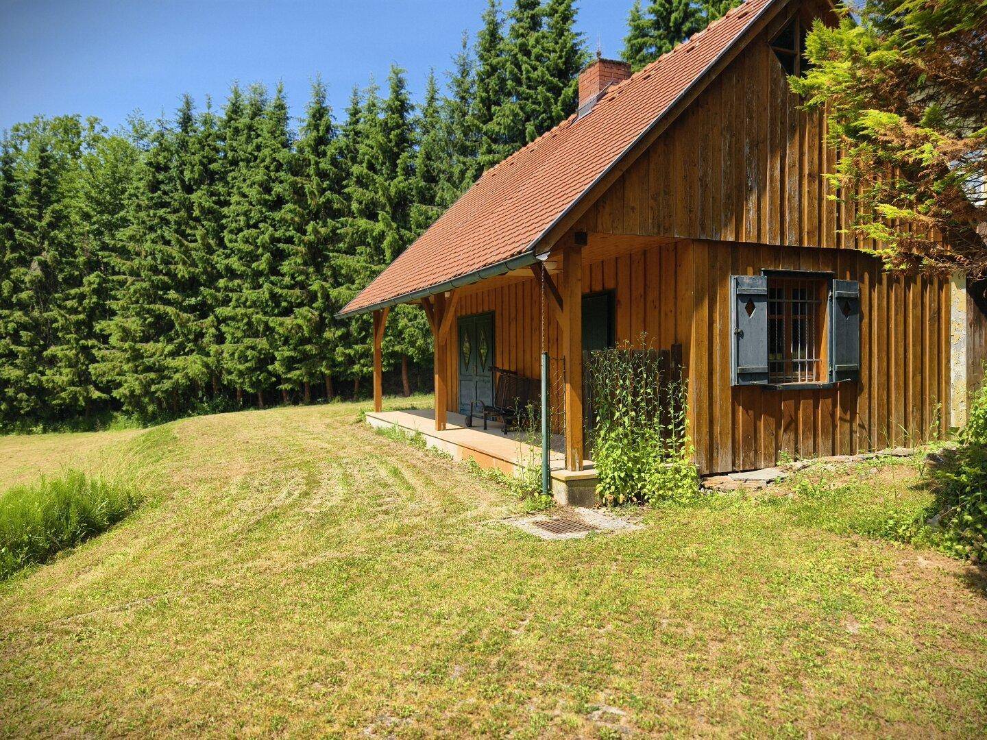 Rustikales Holzhaus mit kleiner Veranda, umgeben von einer grünen Wiese und hohen Nadelbäumen.