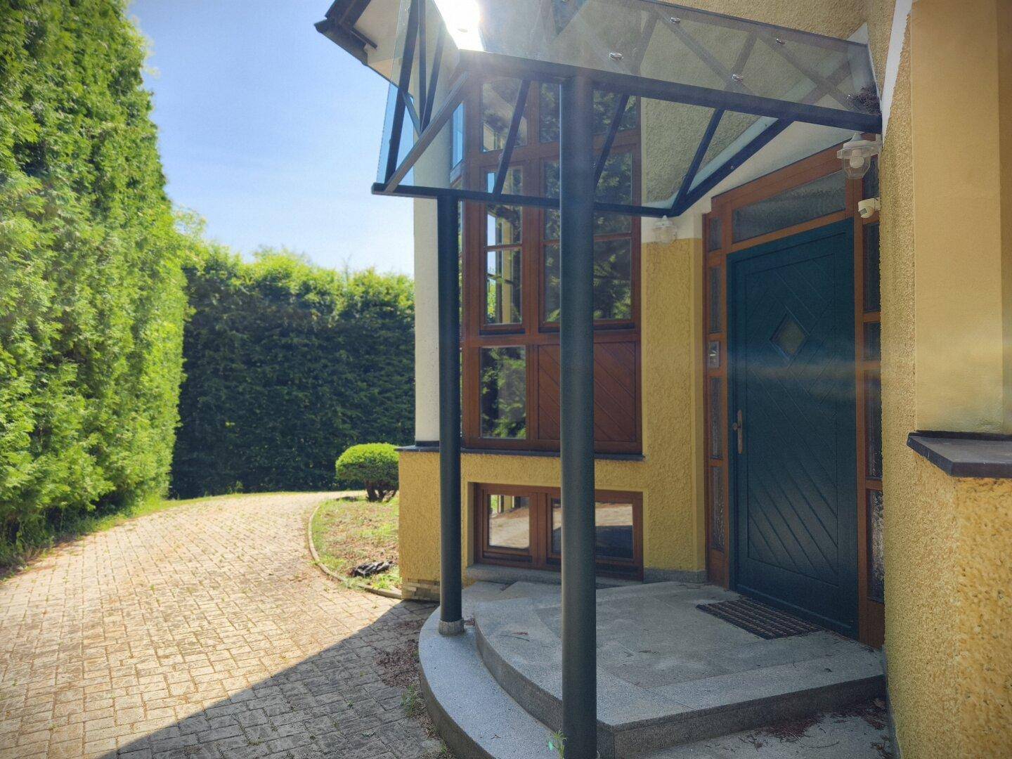 Hauseingang mit Glasvordach und gepflasterter Auffahrt, umgeben von hohen Hecken.