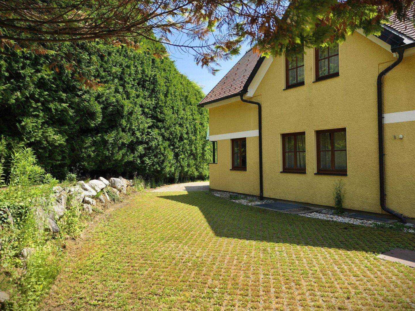 Seitenansicht des Hauses mit grüner Rasenfläche und einer hohen, dichten Hecke entlang der Auffahrt.