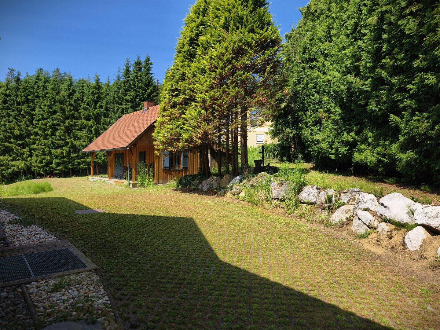 Holzhaus mit Rasenfläche und einem Steingarten, umgeben von hohen Bäumen.