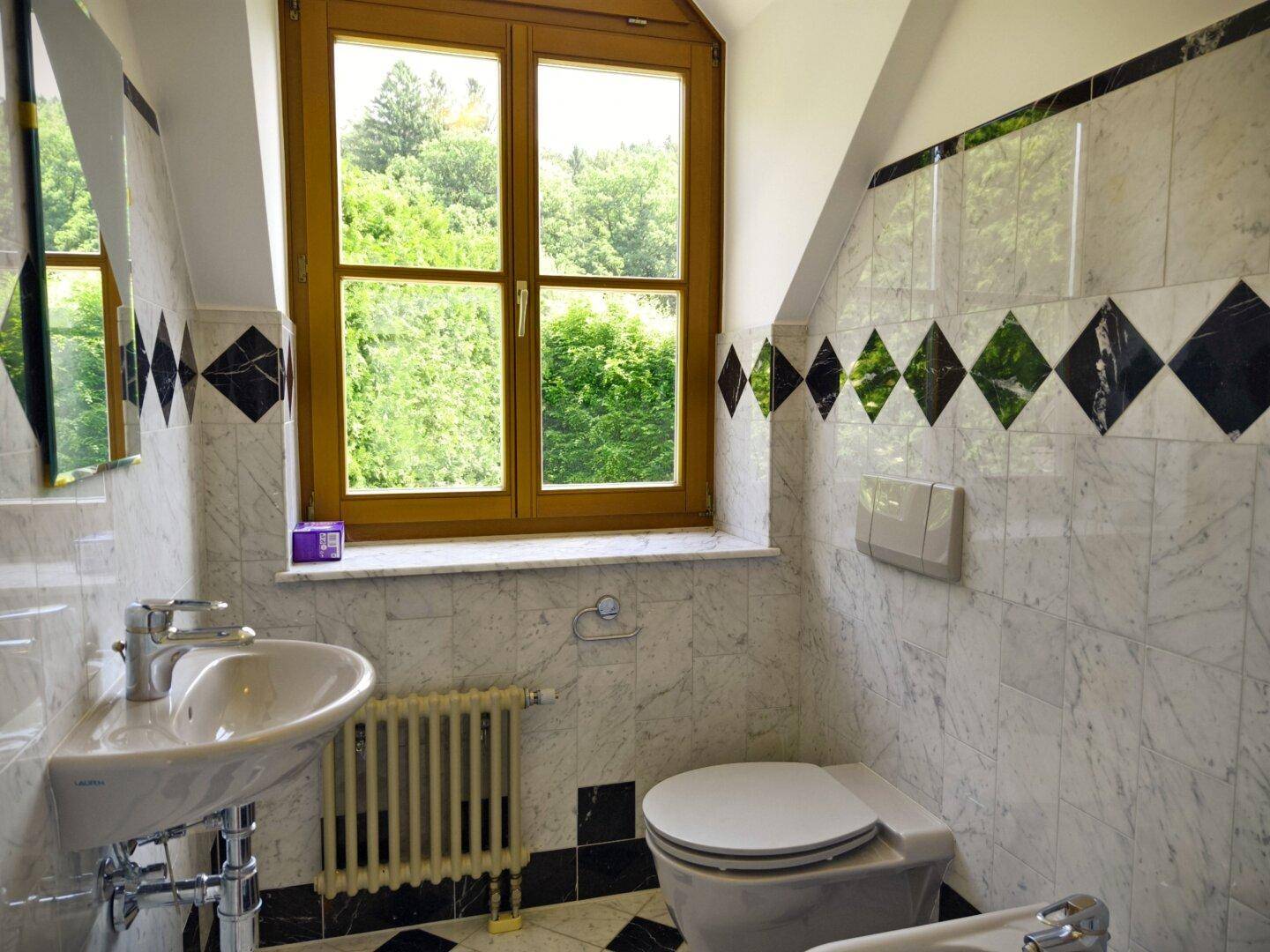 Helles Badezimmer mit Marmorfliesen, Dachschräge und Fenster mit Blick ins Grüne.