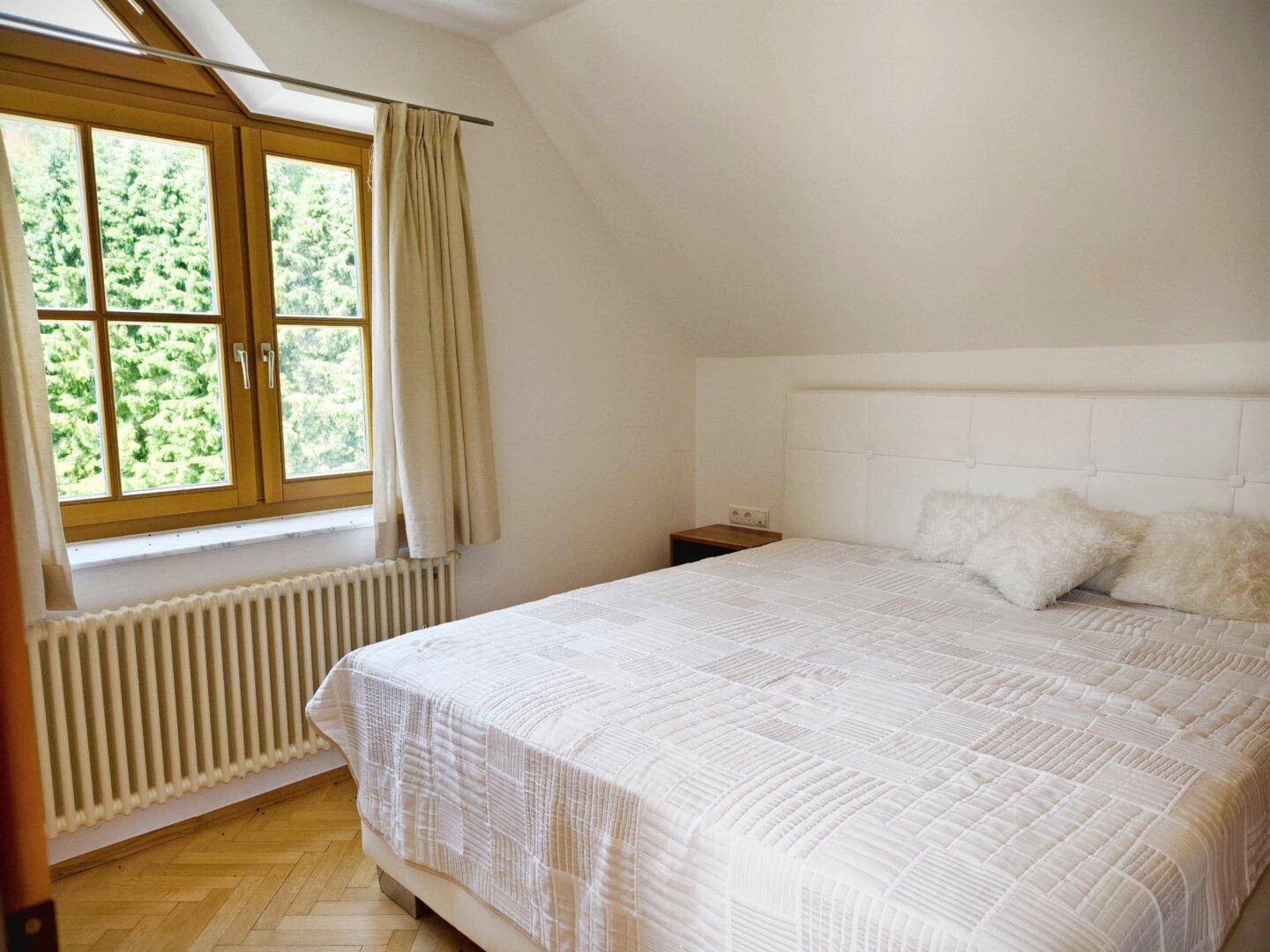 Helles Schlafzimmer mit Doppelbett, Holzparkett und Fenster mit Blick auf die Natur.