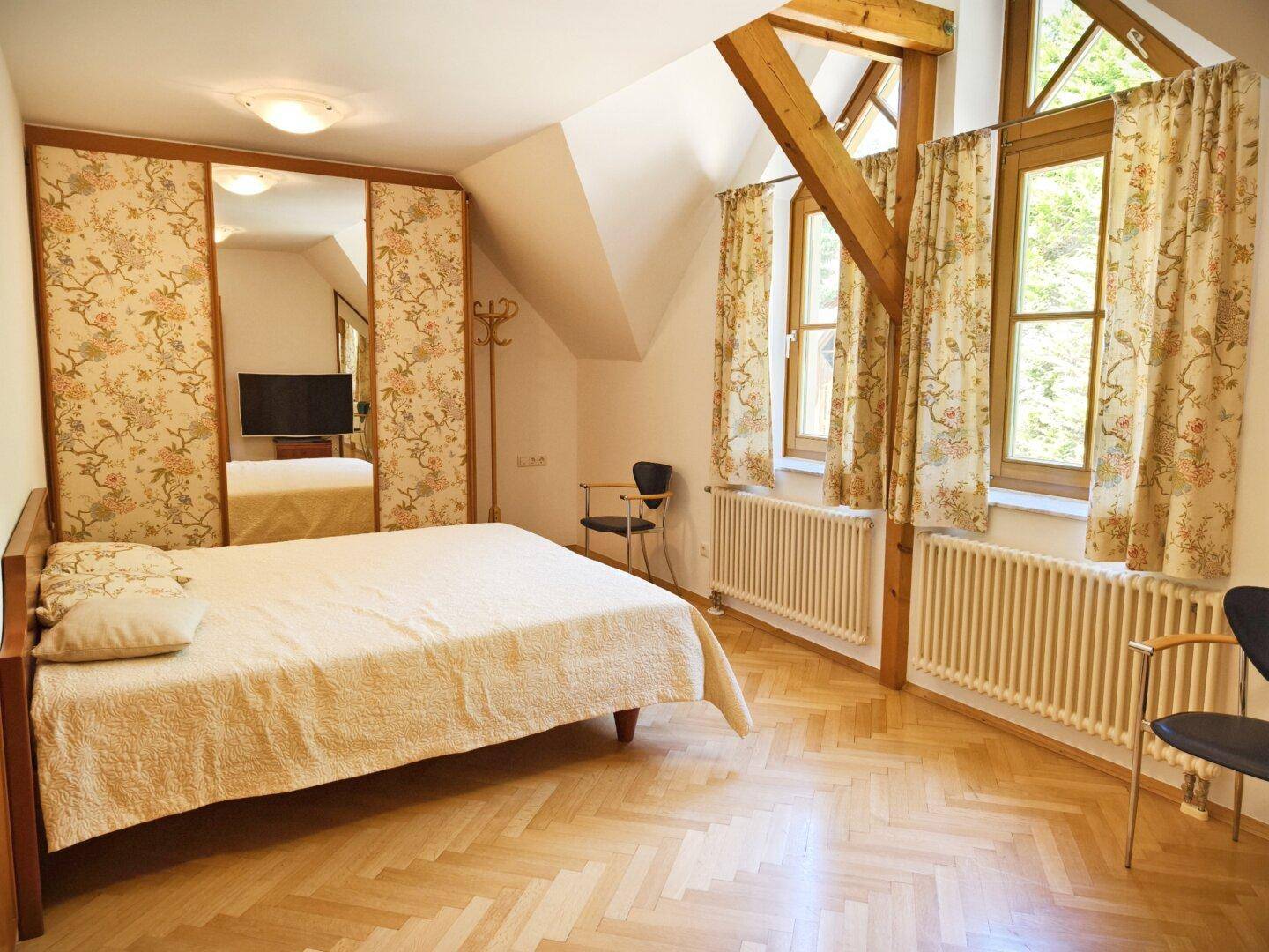 Schlafzimmer mit floral gemustertem Kleiderschrank, Holzparkett und Fenstern mit Blick ins Grüne.