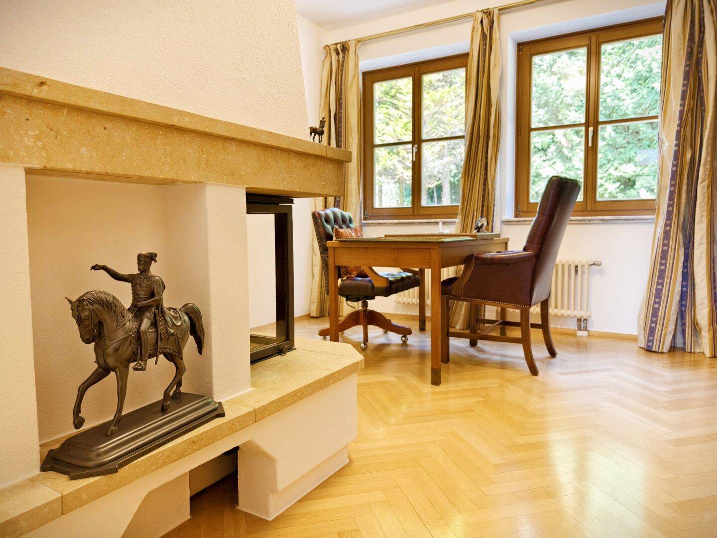 Arbeitszimmer mit Schreibtisch, Kamin und Fenstern mit Blick auf die grüne Umgebung.