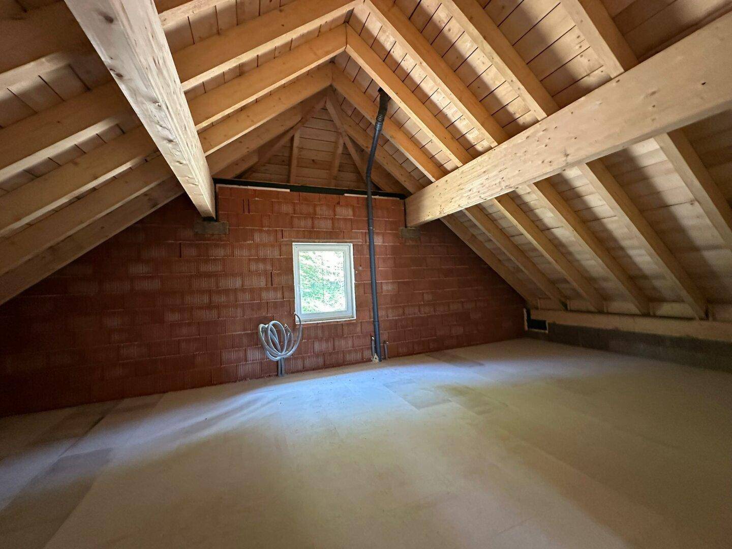Unausgebauter Dachboden mit sichtbaren Holzbalken, Ziegelwänden und einem kleinen Fenster.