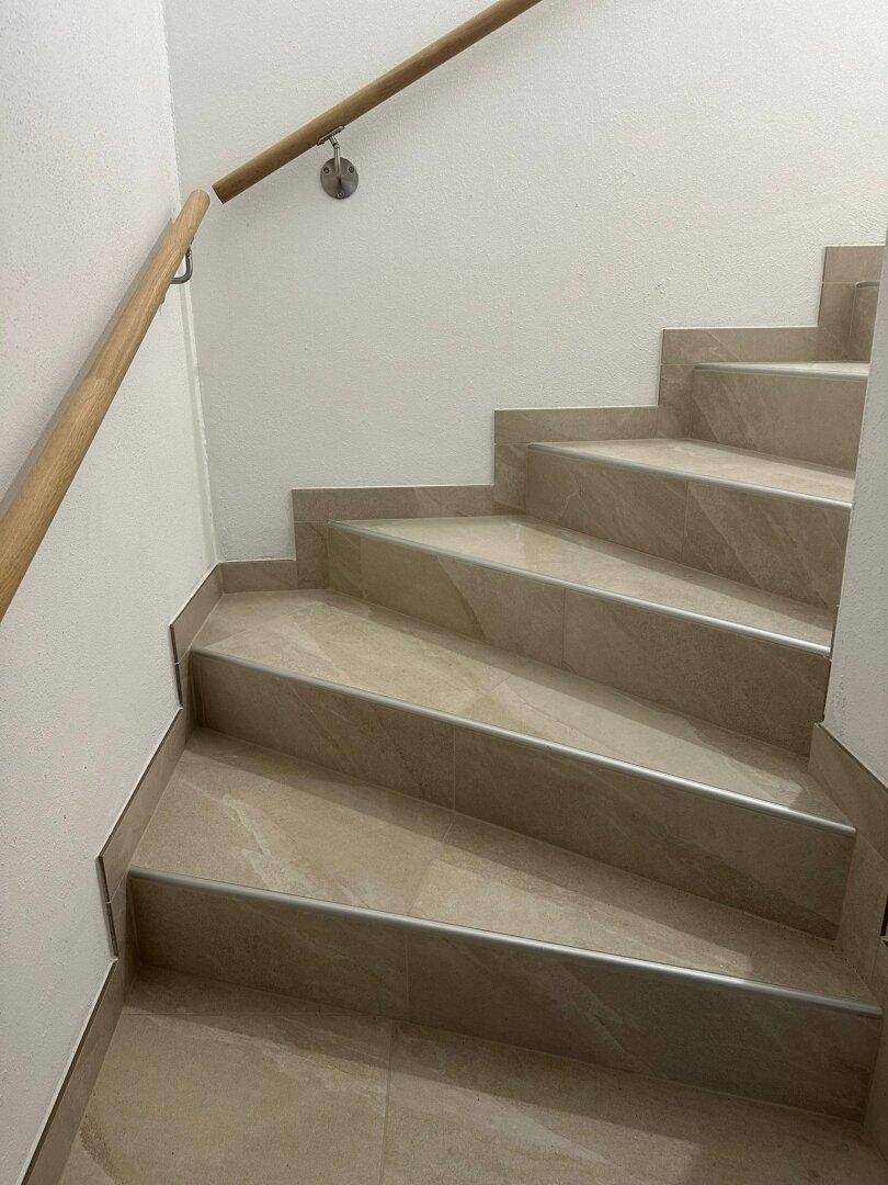 Treppenhaus mit gefliesten Stufen, Holzgeländer und weißen Wänden, das in ein Obergeschoss führt.