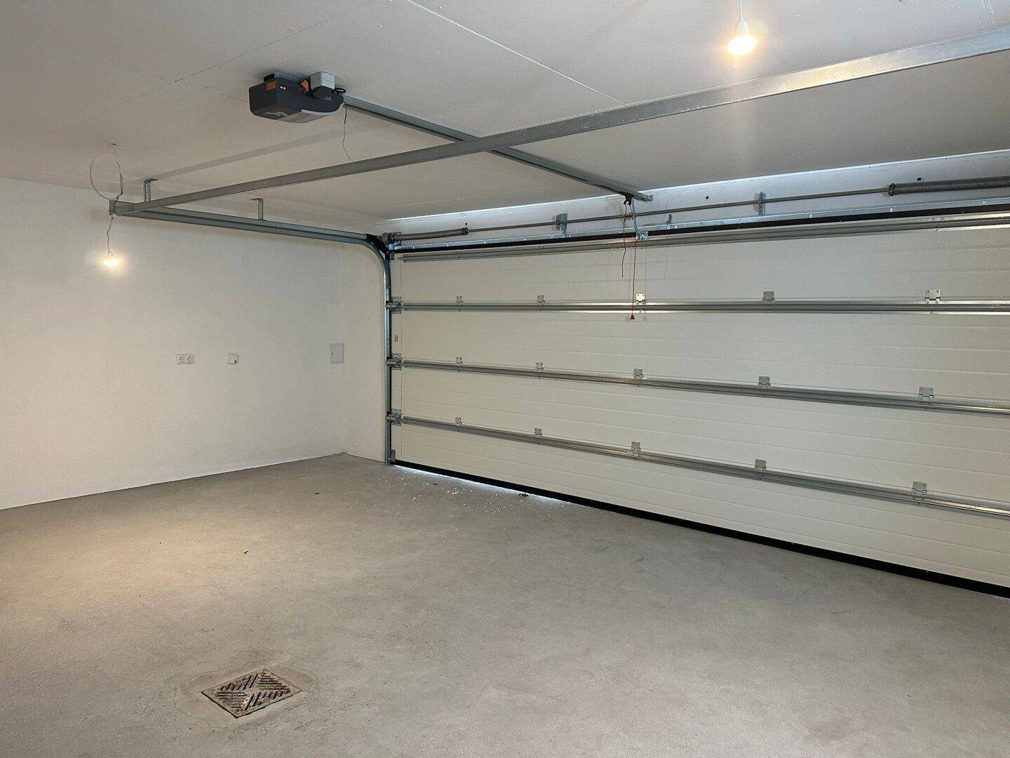 Geräumige Garage mit automatischem Garagentor, Betonboden und Deckenbeleuchtung.