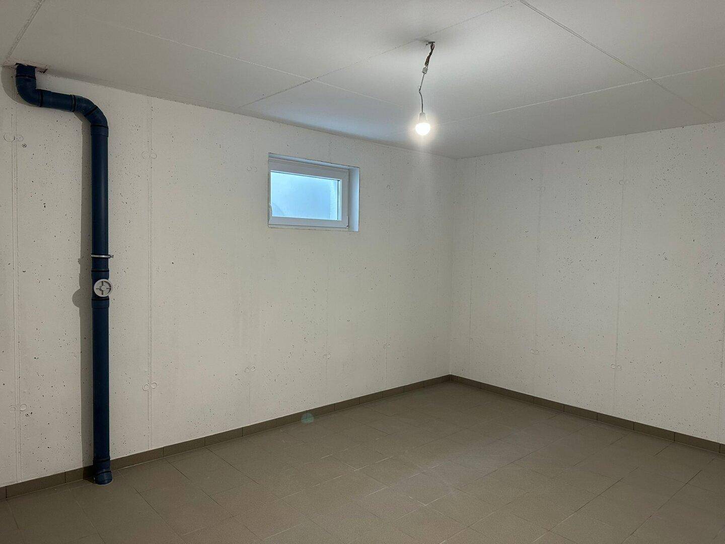 Kellerraum mit kleinem Fenster, grauen Fliesen und sichtbaren blauen Rohren an der Wand.