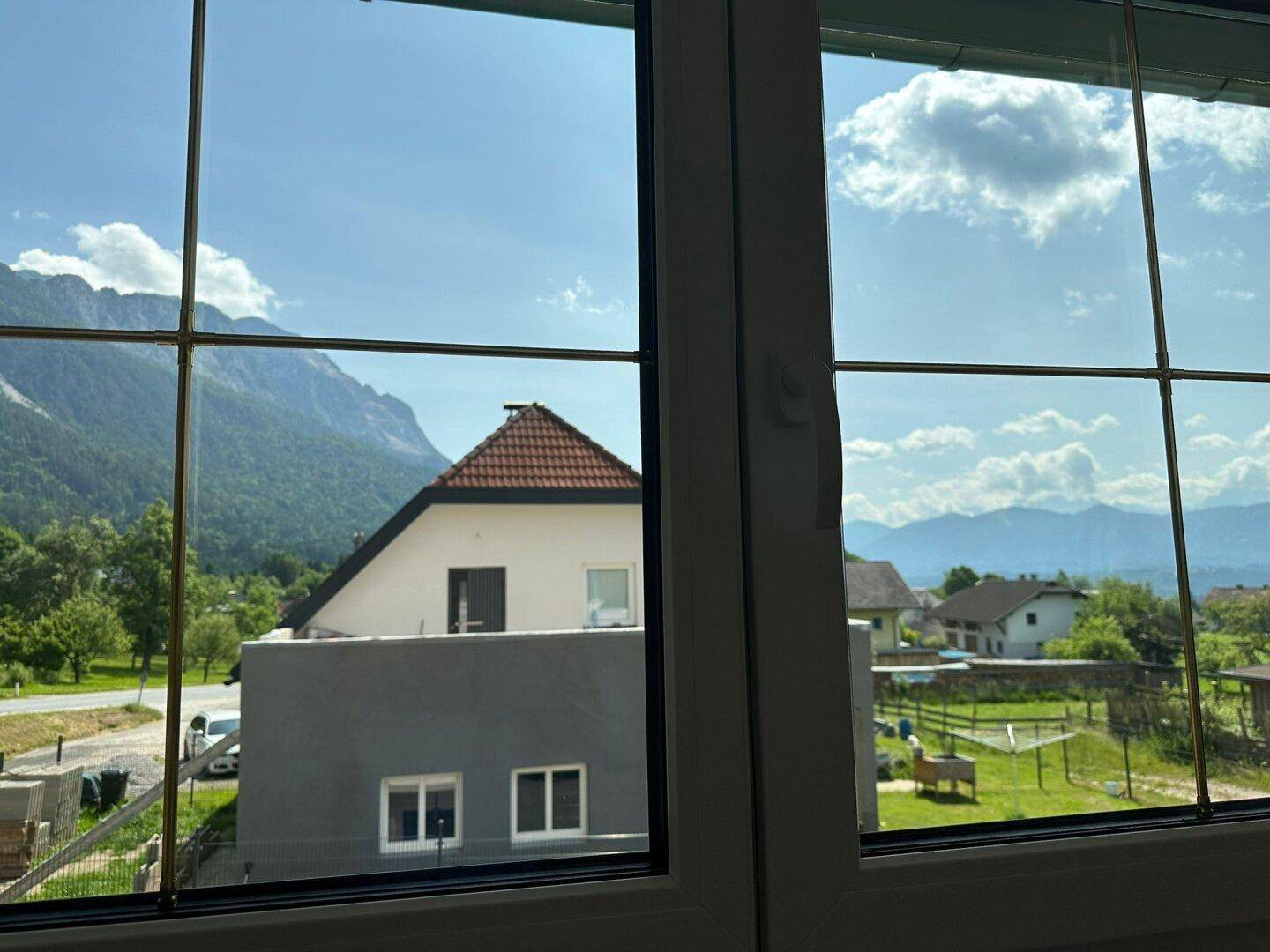 Fensterblick auf ein benachbartes Haus, Berge und grüne Landschaft unter blauem Himmel.