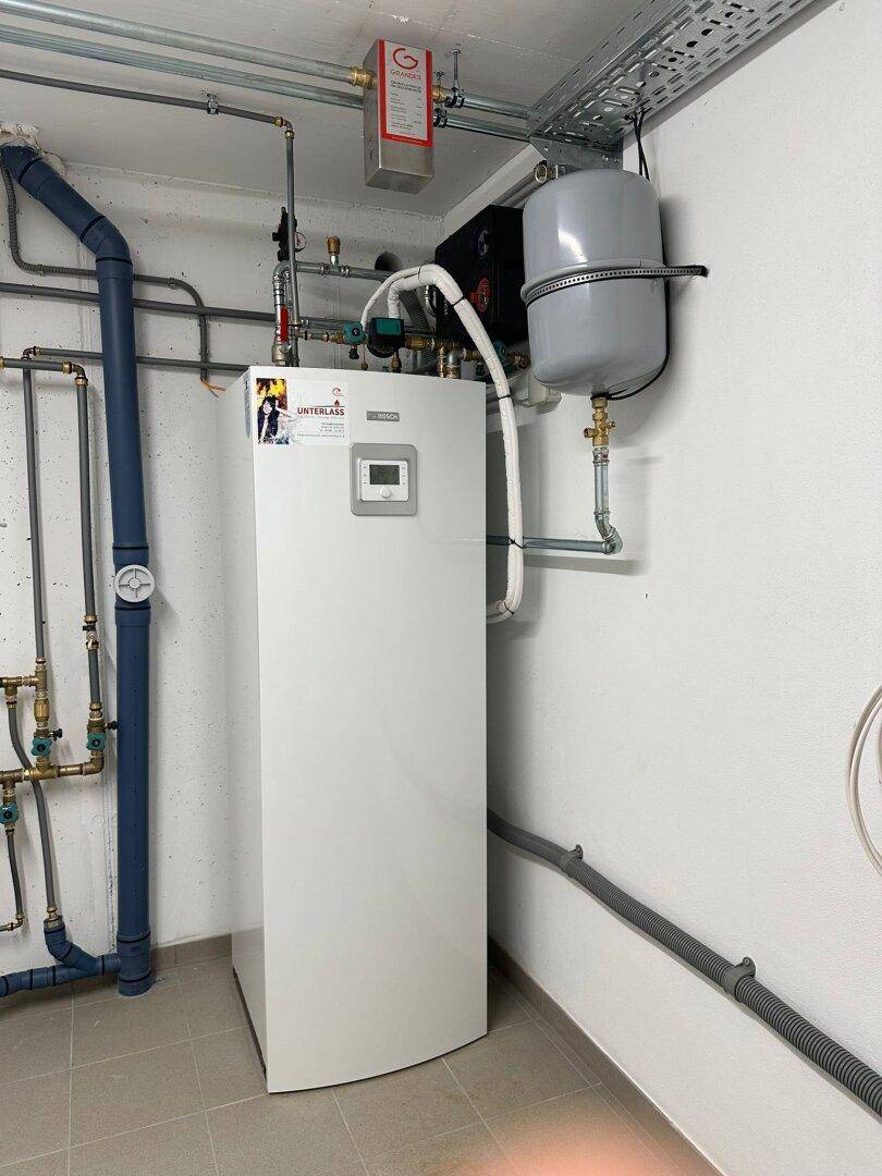 Technikraum mit Wärmepumpe, Warmwasserspeicher und sichtbaren Rohrleitungen an der Wand.