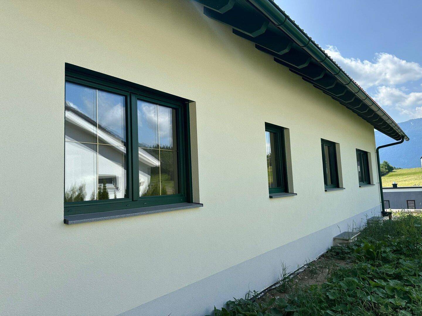 Seitenansicht des Hauses mit grünen Fensterrahmen und heller Fassade unter blauem Himmel.