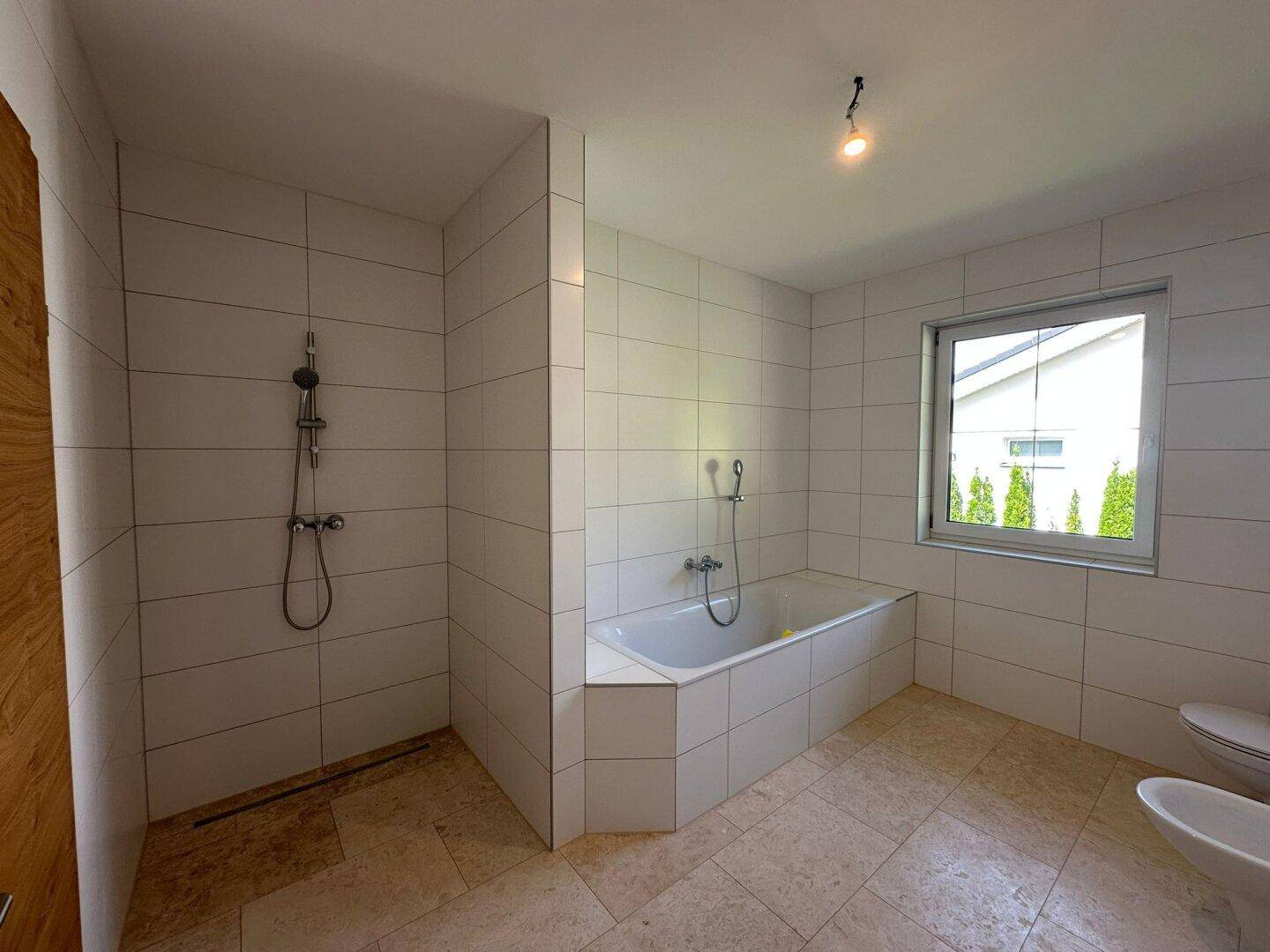 Modernes Badezimmer mit weißen Fliesen, Badewanne, Dusche und Fenster für Tageslicht.