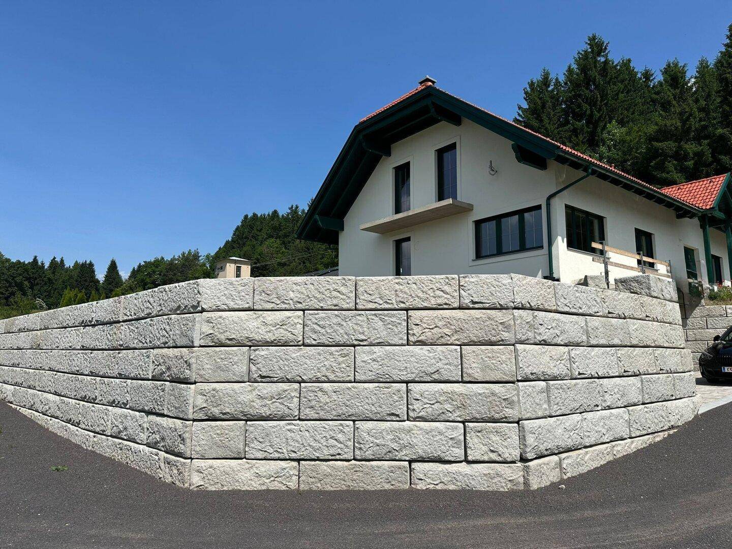 Neubau mit heller Fassade und rotem Dach, umgeben von einer robusten Steinmauer und Bäumen.