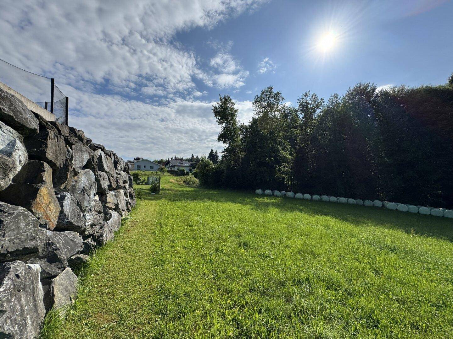 Sonniger Weg entlang einer Steinmauer mit weitem Blick über eine grüne Wiese und Bäume.