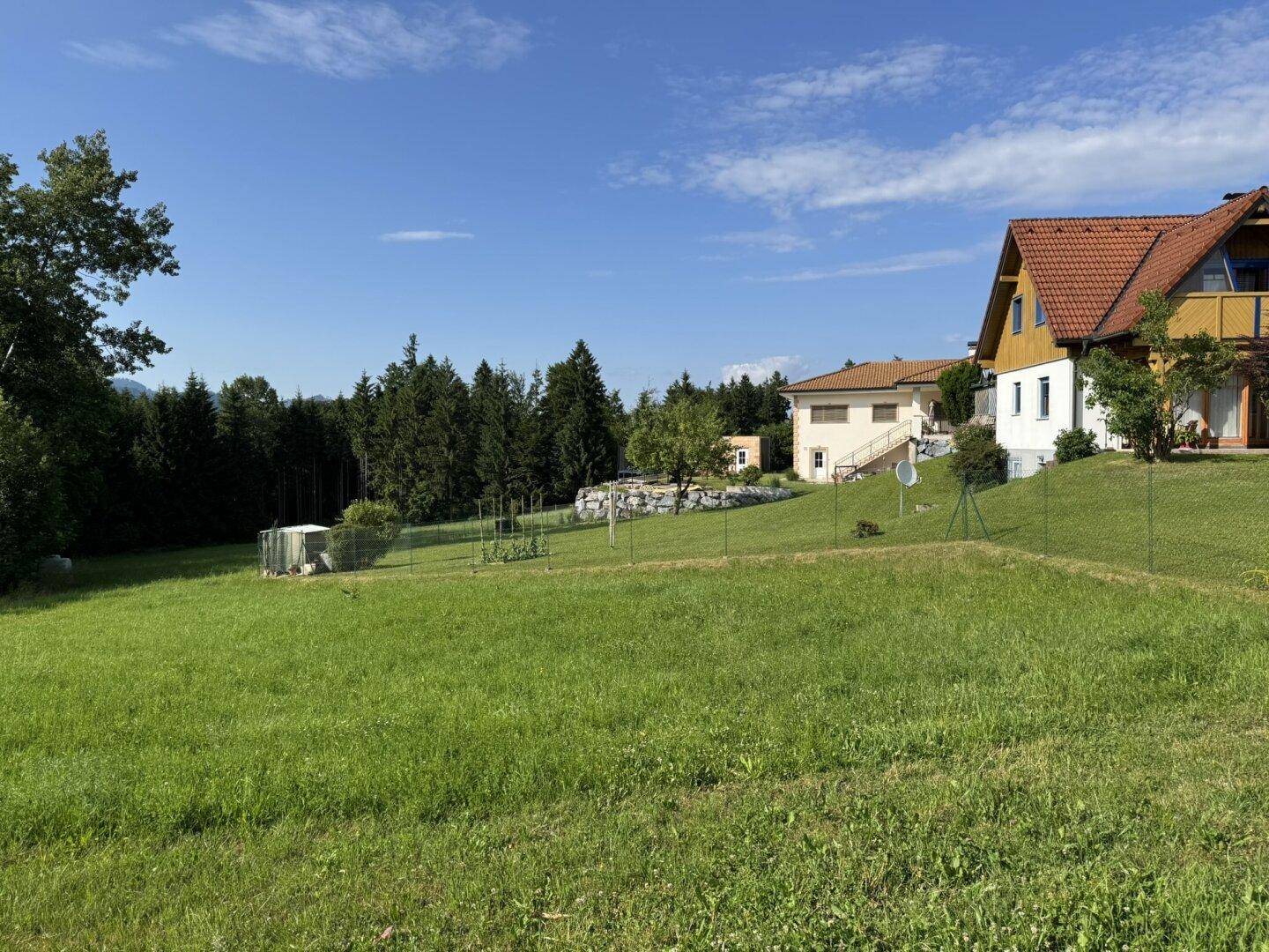 Weitläufige grüne Wiese mit mehreren Wohnhäusern und Bäumen im Hintergrund unter blauem Himmel.