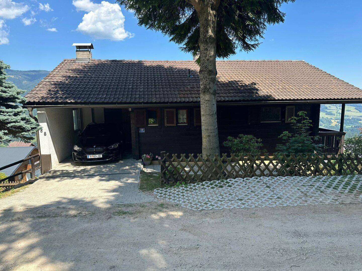 Vorderansicht des Hauses mit integriertem Carport und einem großen Baum im Vorgarten, umgeben von Natur.