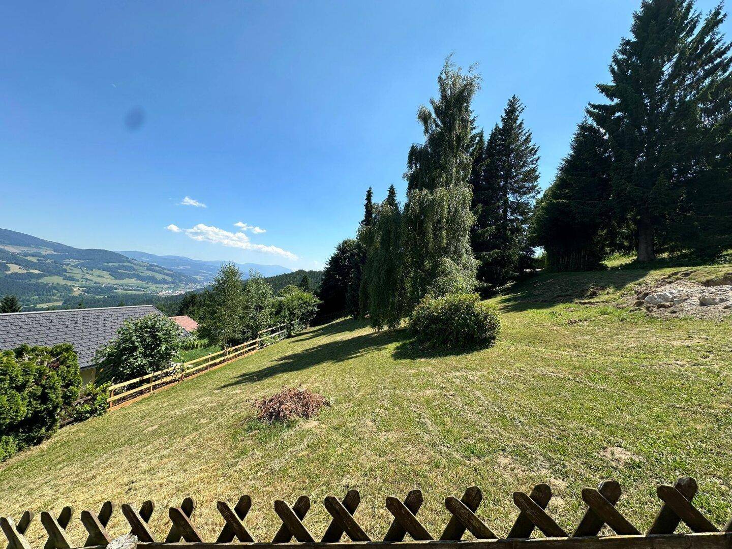 Hanglage mit Rasenfläche und Holzzaun, umgeben von Bäumen und mit Blick auf die Ferne.