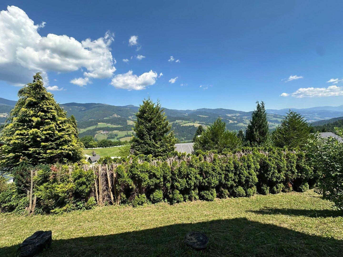 Gartenbereich mit dichter Hecke und weitem Blick auf die grüne Berglandschaft unter strahlend blauem Himmel.