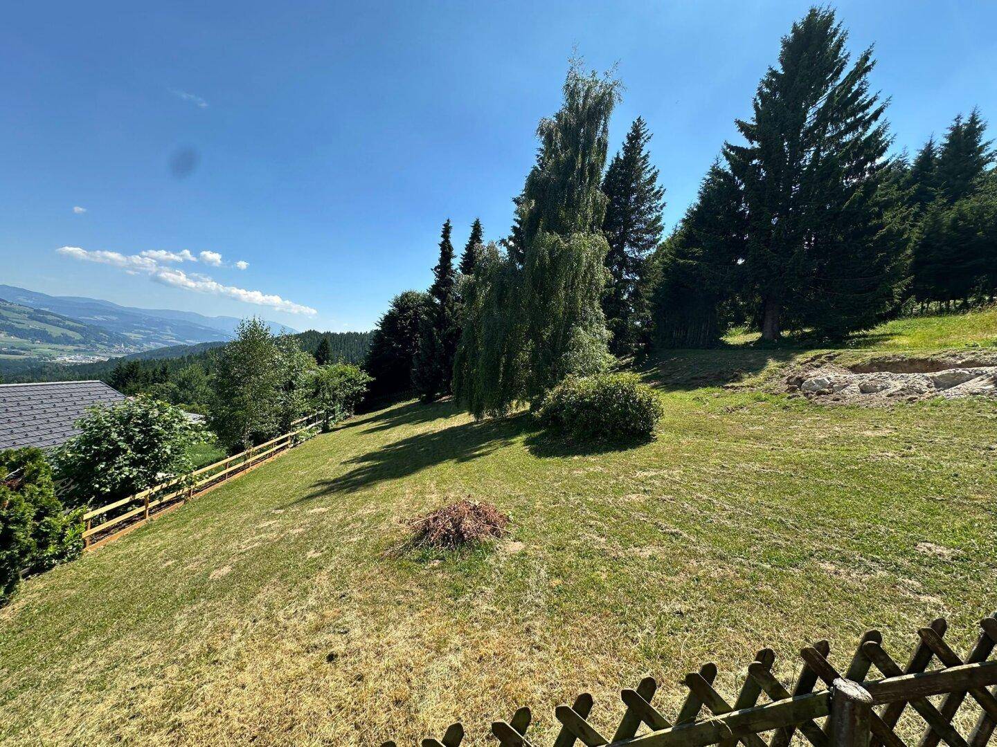 Grüne Hangwiese mit Holzzaun und hohen Bäumen, die einen weiten Blick auf die hügelige Landschaft freigibt.